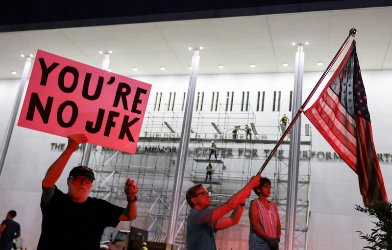 A man waves a U.S. national flag while another holds up a poster, as workers prepare scaffolding preparing ahead of removing lettering from the facade of the John F. Kennedy Memorial Center for the Performing Arts, following a federal judge’s order to remove U.S. President Donald Trump's name from the institution, in Washington, D.C., U.S., June 13, 2026.
